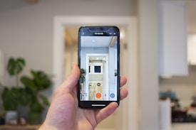 A hand holds a smartphone in a modern, well-lit room. The phone's screen displays a live camera view showing an interior hallway with a plant and bright lighting. In the background, a blurred view of a kitchen is visible with cabinets and appliances.