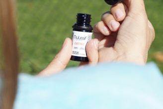 A person's hands are holding a small bottle labeled with 'NuLeaf' and 'Δ8THC'. The person's thumb and index finger are gripping the bottle while the other hand's fingers are visible in the background. The background is blurred and appears to be a grassy area.