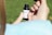 A person's hands are holding a small bottle labeled with 'NuLeaf' and 'Δ8THC'. The person's thumb and index finger are gripping the bottle while the other hand's fingers are visible in the background. The background is blurred and appears to be a grassy area.