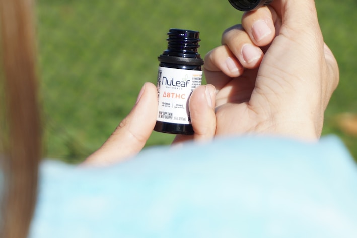 A person's hands are holding a small bottle labeled with 'NuLeaf' and 'Δ8THC'. The person's thumb and index finger are gripping the bottle while the other hand's fingers are visible in the background. The background is blurred and appears to be a grassy area.