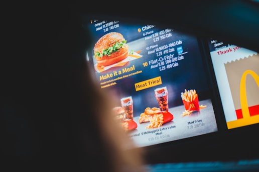 A fast-food menu showcasing various meal options, including a burger, Filet-O-Fish, and chicken nuggets, each with calorie information. The menu features images of meals accompanied by fries and soft drinks, with promotional text, including phrases like 'Make it a Meal' and 'Must Tries!'