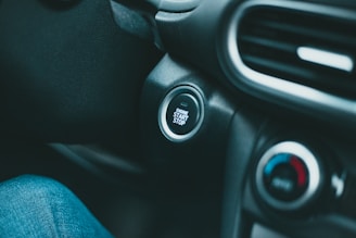 A car interior focusing on the ignition area, featuring a start/stop button surrounded by plastic material. Adjacent controls include a climate control dial showing blue and red indicators. Part of a denim-clad leg is visible in the foreground.