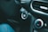 A car interior focusing on the ignition area, featuring a start/stop button surrounded by plastic material. Adjacent controls include a climate control dial showing blue and red indicators. Part of a denim-clad leg is visible in the foreground.