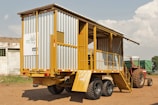 A mobile structure resembling a trailer is parked on a dirt ground. It has a metal frame with yellow and gray siding, featuring stairs leading to an open section. In the background, a red tractor is partially visible. The sky is partly cloudy, and a small building can be seen on the left.