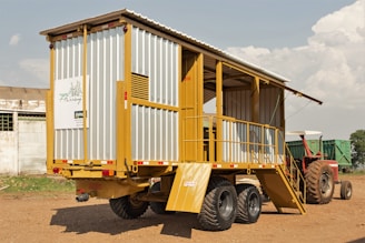 A mobile structure resembling a trailer is parked on a dirt ground. It has a metal frame with yellow and gray siding, featuring stairs leading to an open section. In the background, a red tractor is partially visible. The sky is partly cloudy, and a small building can be seen on the left.