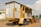 A mobile structure resembling a trailer is parked on a dirt ground. It has a metal frame with yellow and gray siding, featuring stairs leading to an open section. In the background, a red tractor is partially visible. The sky is partly cloudy, and a small building can be seen on the left.