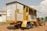 A mobile structure resembling a trailer is parked on a dirt ground. It has a metal frame with yellow and gray siding, featuring stairs leading to an open section. In the background, a red tractor is partially visible. The sky is partly cloudy, and a small building can be seen on the left.