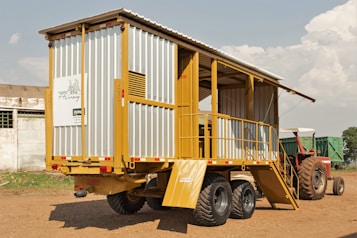 A mobile structure resembling a trailer is parked on a dirt ground. It has a metal frame with yellow and gray siding, featuring stairs leading to an open section. In the background, a red tractor is partially visible. The sky is partly cloudy, and a small building can be seen on the left.