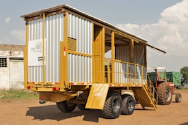 A mobile structure resembling a trailer is parked on a dirt ground. It has a metal frame with yellow and gray siding, featuring stairs leading to an open section. In the background, a red tractor is partially visible. The sky is partly cloudy, and a small building can be seen on the left.