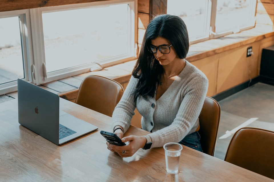 Woman typing on a phone while sitting at a table with a plastic cup of water and an open laptop.