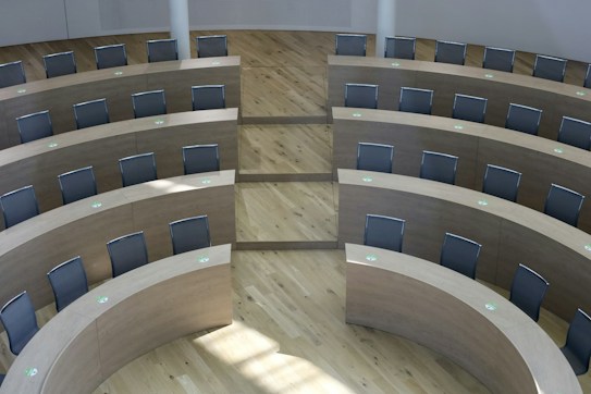 An empty, modern lecture hall featuring curved, tiered rows of wooden desks and black chairs. The floor is made of light wood, and the room is well-lit with natural light coming in from the left side. Each desk has a green marking, possibly indicating social distancing measures.