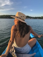 A woman wearing a straw hat is sitting in a kayak on a calm lake. Her back is towards the camera, showcasing a backless top and denim shorts. The sun is setting, casting a warm glow on her face and the surrounding water. In the distance, a shoreline with trees and houses can be seen.