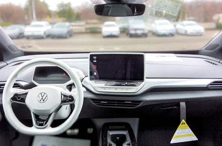 A car interior featuring a modern and minimalistic dashboard design. A large touchscreen display is centrally located with tactile buttons below it. The steering wheel is branded with a Volkswagen logo and has several control buttons. Outside, several parked cars are visible through the windshield.