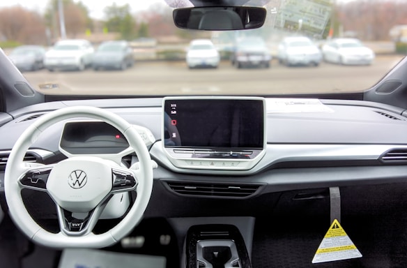 A car interior featuring a modern and minimalistic dashboard design. A large touchscreen display is centrally located with tactile buttons below it. The steering wheel is branded with a Volkswagen logo and has several control buttons. Outside, several parked cars are visible through the windshield.