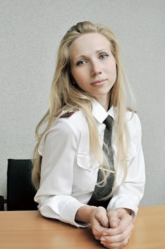 A young woman with long blonde hair is wearing a white uniform shirt with two pockets and epaulets, along with a black tie. She is sitting at a wooden table, resting her hands in front of her. The background is a plain light gray wall.