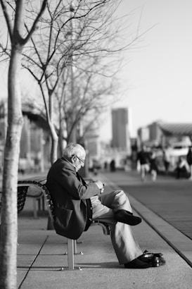 An elderly man sits alone on a bench along a sidewalk, engrossed in a small device, possibly a phone. Leafless trees line the walkway, and a cityscape is visible in the blurred background. The scene has a serene and contemplative atmosphere.