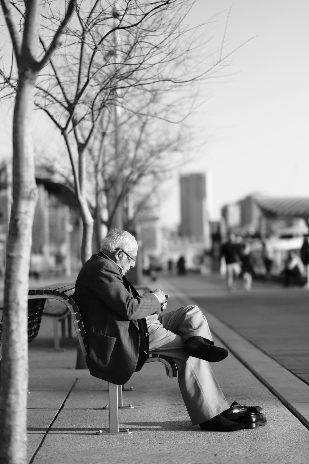 grayscale photo of man sitting on bench near bare trees
