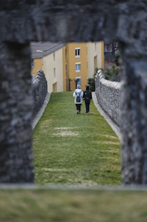 Two people with backpacks are walking down a path flanked by stone walls, heading towards yellow buildings in the background. The photo is framed by an archway made of stone at the foreground.