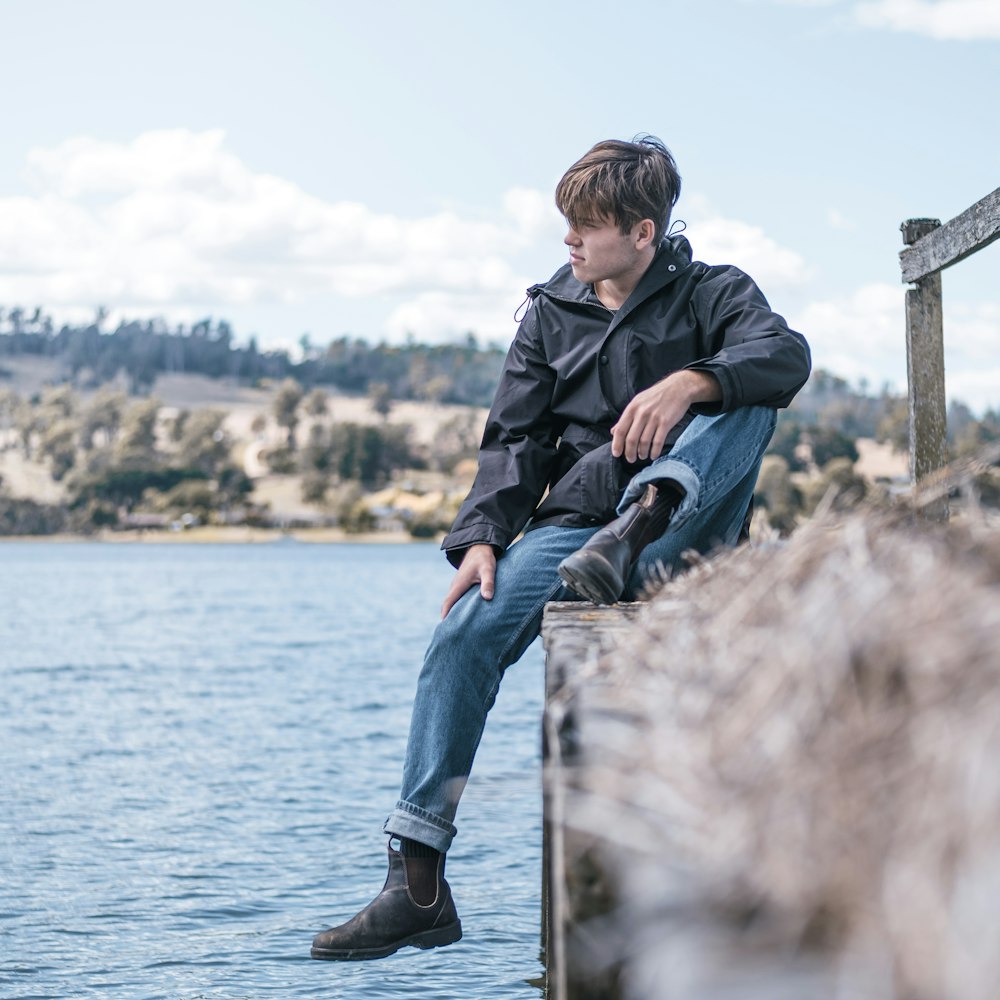 man in black jacket and blue denim jeans sitting on wooden post near body of water