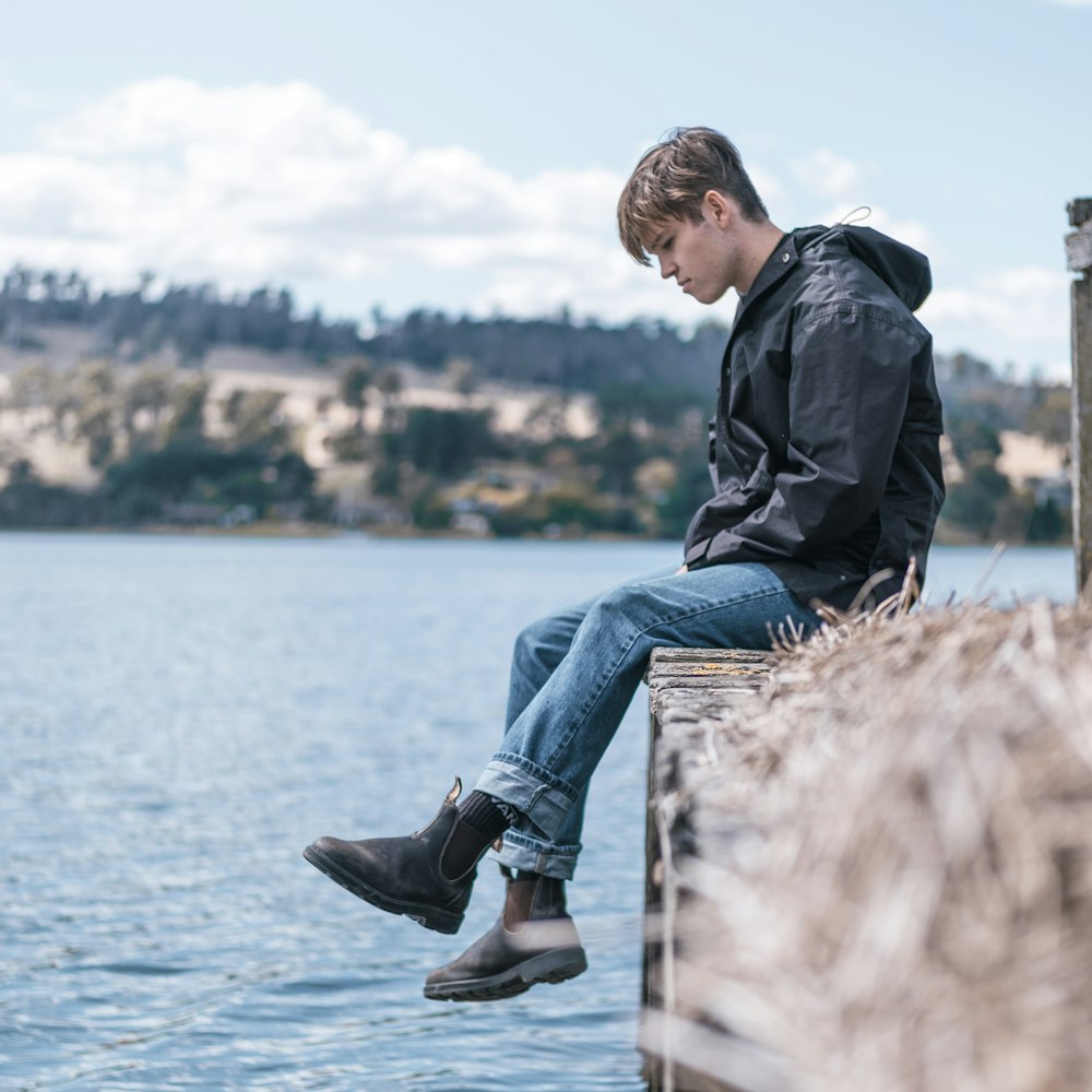 man in black jacket and blue denim jeans sitting on brown wooden log during daytime