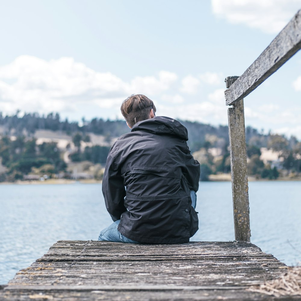 man in black jacket sitting on wooden dock during daytime