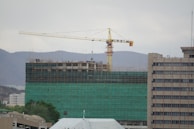 A construction site with a tall building covered in green netting and scaffolding, with a large yellow crane towering above it. Several workers are visible on the top floor. In the background, there are hills and other buildings.
