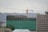 A construction site with a tall building covered in green netting and scaffolding, with a large yellow crane towering above it. Several workers are visible on the top floor. In the background, there are hills and other buildings.