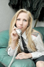 A person with long blonde hair is sitting on a teal leather couch, holding a phone with a coiled cord. The individual is dressed in a white shirt with epaulettes and a black tie, suggesting a professional or uniformed attire. In the background, there are dark green plant leaves.