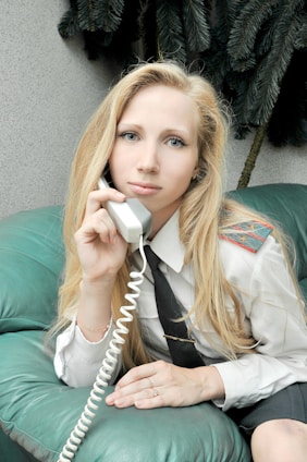 A person with long blonde hair is sitting on a teal leather couch, holding a phone with a coiled cord. The individual is dressed in a white shirt with epaulettes and a black tie, suggesting a professional or uniformed attire. In the background, there are dark green plant leaves.