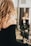 A person with long, wavy blond hair wears a black off-shoulder top, standing in front of a mirror. In the background, another person with a face mask is seated, wearing a black salon cape in a salon setting.