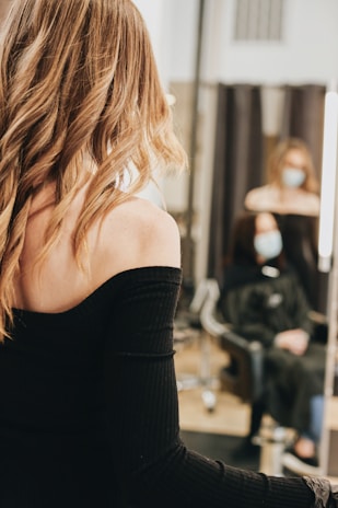 A person with long, wavy blond hair wears a black off-shoulder top, standing in front of a mirror. In the background, another person with a face mask is seated, wearing a black salon cape in a salon setting.