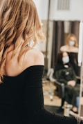 A person with long, wavy blond hair wears a black off-shoulder top, standing in front of a mirror. In the background, another person with a face mask is seated, wearing a black salon cape in a salon setting.