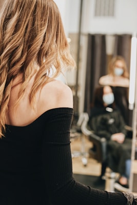 A person with long, wavy blond hair wears a black off-shoulder top, standing in front of a mirror. In the background, another person with a face mask is seated, wearing a black salon cape in a salon setting.