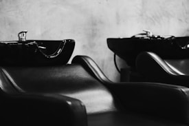 Black and white photograph featuring sleek, modern salon chairs with attached black washing basins. The chairs are upholstered in smooth black material and positioned against a plain, light-textured wall.
