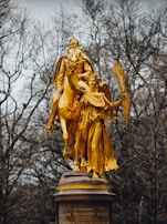 A striking golden statue of a historical figure on horseback, accompanied by an angelic figure holding a palm branch. They are positioned on a stone pedestal against a backdrop of leafless trees.