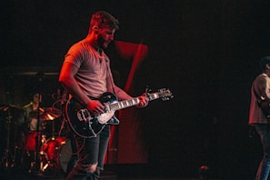 A musician passionately plays an electric guitar on stage, surrounded by dim lighting that emphasizes a warm, moody atmosphere. In the background, another musician is seen playing drums, adding to the live concert setting.