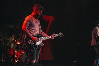 A musician passionately plays an electric guitar on stage, surrounded by dim lighting that emphasizes a warm, moody atmosphere. In the background, another musician is seen playing drums, adding to the live concert setting.