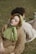 A woman with closed eyes lies on a man's back in a grassy outdoor setting. She is wearing a light green headscarf and a brown sweater, while the man is dressed in beige attire. The setting appears calm and serene, with soft lighting emphasizing a peaceful moment.