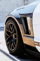 A close-up of the rear wheel and fender of a sleek car with a carbon fiber texture. The vehicle has a low profile and features glossy black rims with a stylish pattern. Sunlight casts sharp shadows on the ground, highlighting the polished finish.