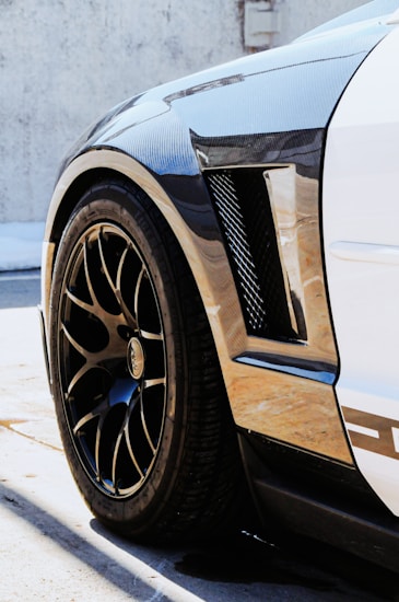 A close-up of the rear wheel and fender of a sleek car with a carbon fiber texture. The vehicle has a low profile and features glossy black rims with a stylish pattern. Sunlight casts sharp shadows on the ground, highlighting the polished finish.