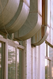Large, old industrial air conditioning or ventilation ducts on the exterior of a building. The ducts are metallic and show signs of rust and wear. There are large windows along the brick wall of the building, reflecting the surroundings.