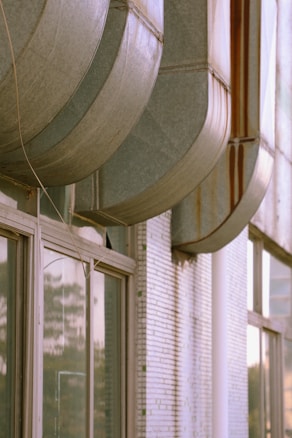 Large, old industrial air conditioning or ventilation ducts on the exterior of a building. The ducts are metallic and show signs of rust and wear. There are large windows along the brick wall of the building, reflecting the surroundings.