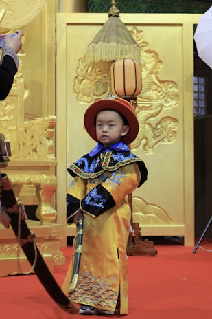 A young child dressed in traditional, elaborate clothing with gold and blue embroidery stands on a red carpet in front of an ornate, gold backdrop. The clothing includes a long robe and matching hat. The background features intricate patterns and designs, including what appears to be a large decorative panel.