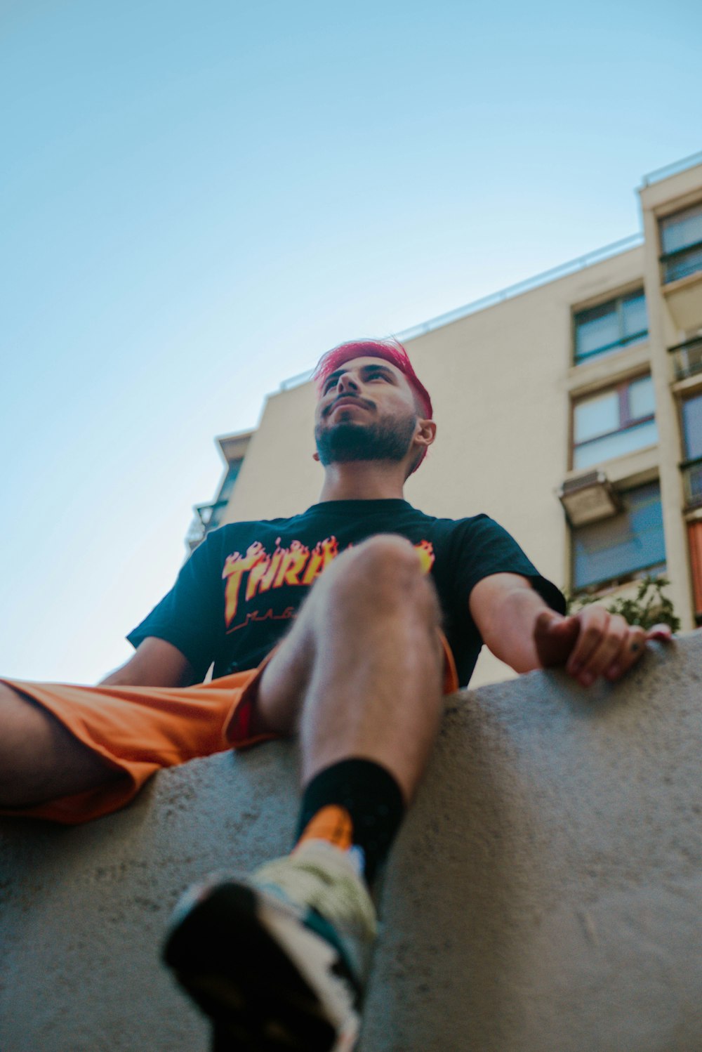 man in black crew neck t-shirt and orange shorts sitting on concrete wall during daytime