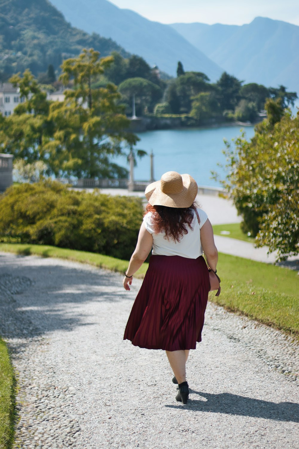 woman in red dress wearing brown sun hat walking on gray asphalt road during daytime