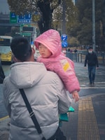 A person wearing a light-colored jacket is carrying a child in a pink hooded jacket. The child appears to be looking at the camera with a curious expression. In the background, there are street signs, trees, a yellow bus, and people walking, some wearing masks.