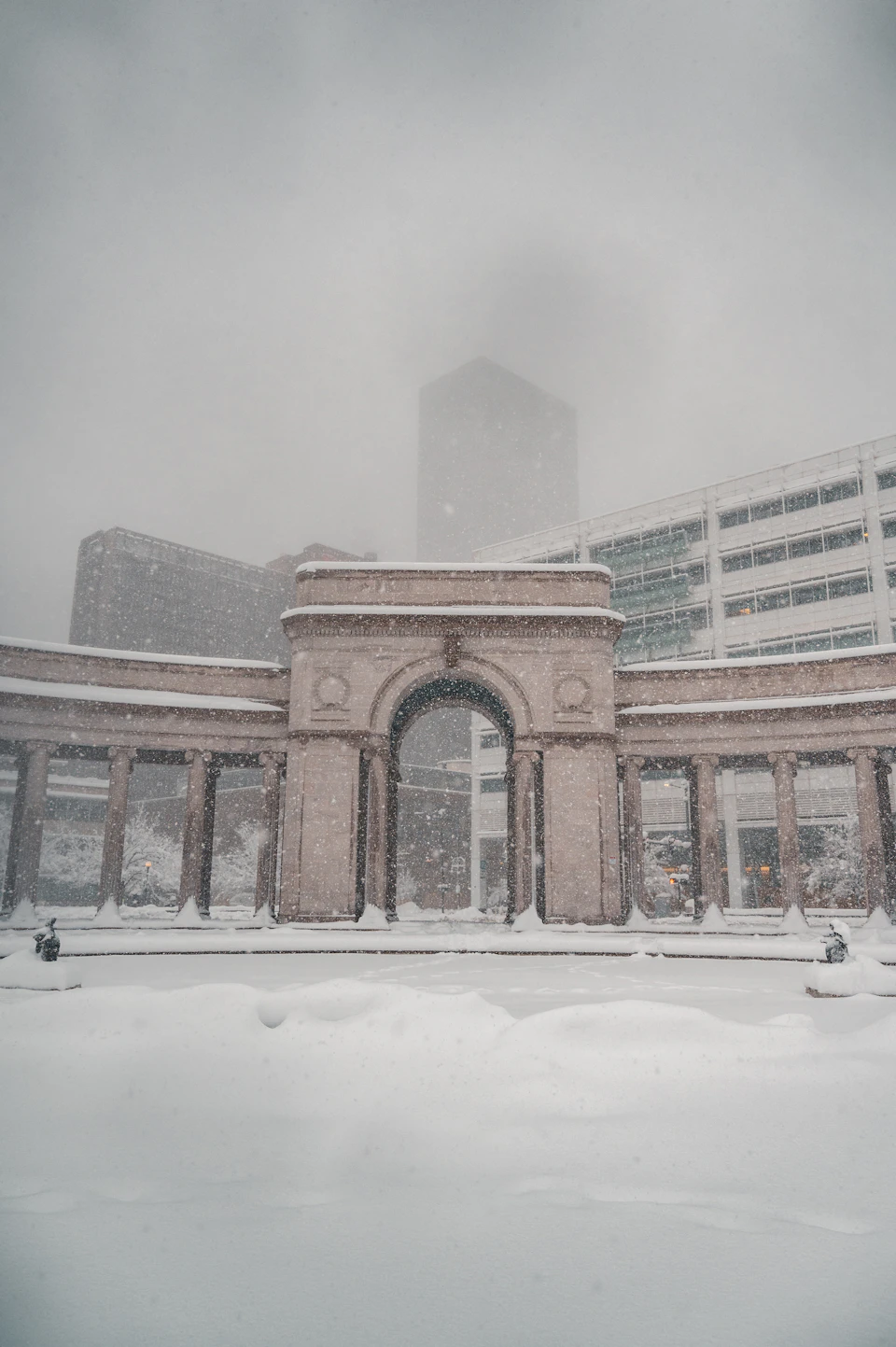The image depicts a large, ornate archway constructed from stone, standing tall against an overcast sky. The archway is adorned with intricate carvings and features two columns on either side, adding to its grandeur. The surrounding area is blanketed in snow, creating a serene winter landscape that contrasts with the architectural marvel of the archway.

In the background, there are several buildings visible, including one large building on the left side of the image and another smaller structure on the right side. The sky above is filled with clouds, suggesting an overcast day.