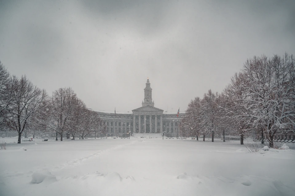 The image depicts a large, white building with columns and a dome, standing out against the snowy landscape. The building is surrounded by trees on all sides, creating a sense of isolation. The sky above is overcast, casting a soft light over the scene. The snow-covered ground reflects the muted colors of the surroundings, enhancing the wintry atmosphere.