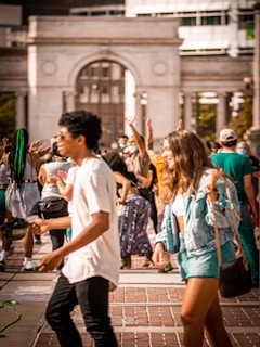 A busy outdoor scene with people walking and gathering in front of a large classical building. Individuals are dressed in casual attire, some with hands raised. The atmosphere suggests a lively and engaging public event.