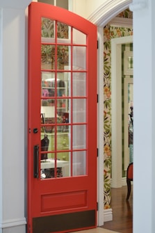 A vibrant red door with glass panels is partially open, revealing an interior with floral wallpaper and a room beyond. The room has white and green elements, including a visible piece of furniture. The image shows a mix of contemporary and classic design elements, with ornate wallpaper and traditional door styling.